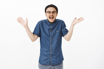 Studio shot of excited and impressed man cannot believe in own luck shrugging with palms spread aside retelling amazing story happened to him being under great influence and thrill over gray wall