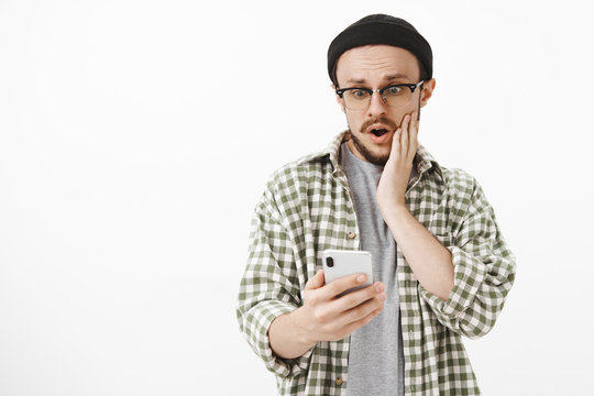 Unpopular guy being shocked receiving likes under latest post in social network gasping with opened mouth holding hand on cheek staring surprised and amazed at smartphone screen over white wall