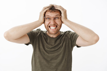 Fototapeta premium Troubled and perplexed guy needs vacation. Portrait of distressed annoyed young male office worker in green t-shirt and glasses clenching teeth holding hands on head in hopeless pose having crappy day