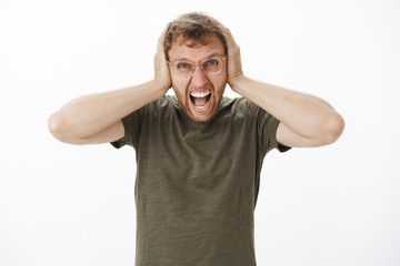 Guy lose temper being pissed with loud noises at night yelling out loud with angry outrage expression covering ears with palms on head being in rage over white background in casual dark-green t-shirt