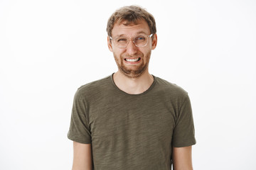 Fototapeta premium Guy squinting and making funny face eating sour lemon frowning and wrinkling nose standing disgusted and dissatisfied over white background in transparent glasses and green t-shirt