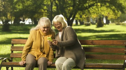 Elderly woman showing family photos on phone to friend, sitting on bench in park - Powered by Adobe