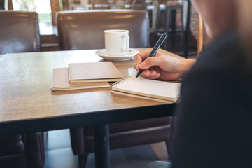 Closeup image of a woman writing on blank notebook with coffee cup on table in cafe