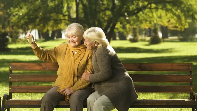 Two old ladies taking selfie on mobile phone sitting on bench in park technology