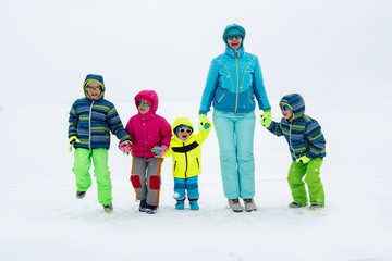 Fototapeta na wymiar Happy young mother in blue ski suit wearing sunglasses with funny children in bright winter clothes jumping for joy on shore of icy lake. Wonderful winter vacation for whole family. Kids fun together