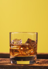 glass of whiskey and ice on wooden table yellow background