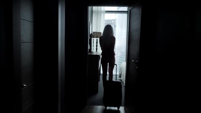 Young blond businesswoman arrives in a hotel room with black suitcase. Woman in red coral business suit. Young girl enters the room holding the suitcase by the handle