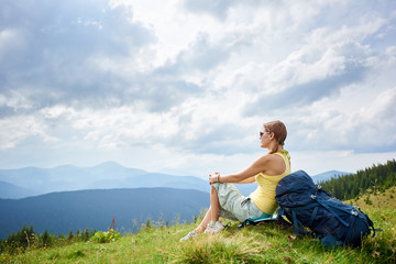 Fototapeta na wymiar Side view of attractive woman hiker resting on grassy hill with backpack, wearing sunglasses. Female tourist enjoying summer cloudy day in the Carpathian mountains. Outdoor activity, tourism concept