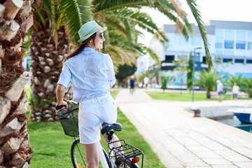 Back view of young happy woman in sunglasses, white shorts, blouse and hat riding a bicycle on green palm tree background on bright sunny day. Active lifestyle and vacations concept.