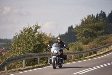 Smiling motorcyclist in black leather outfit and sunglasses riding cruiser motorbike with stretched...
