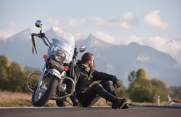 Handsome motorcyclist in black leather clothing sitting at cruiser powerful shiny motorbike on roadside on blurred copy space background of distant mountain peaks.
