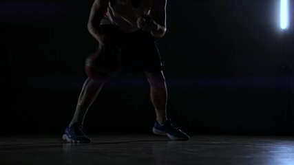 A man with a basketball on a dark basketball court against the backdrop of a basketball ring in the smoke shows dribbling skills illuminated by three lanterns in backlight