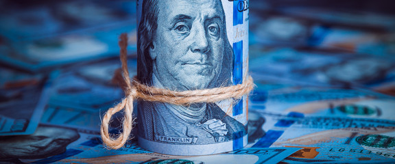 A roll of dollars with coins on the background of scattered one hundred dollar bills in blue light.