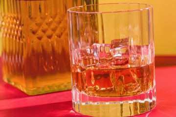 glass of whiskey and ice on red and yellow background with decanter closeup