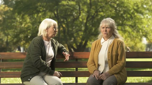 Two grumpy old ladies judging passerby people, sitting on bench in park, pension
