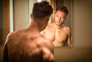 Fototapeta na wymiar Shirtless handsome young man examining his face in bathroom mirror in the morning