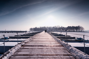 Frozen pier on a winter lake. Two people walk on a snow-covered bridge in the sun