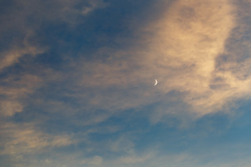 A growing moon in the blue sky among the clouds illuminated by the sun at sunset