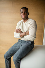 Portrait of young African American male student wearing white pullover and formal trousers, sitting inside university campus, looking at camera with pleased smile, happy to pass exams