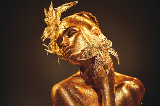 Fototapeta Fashion gold model woman with bright golden sparkles on skin posing, fantasy flower. Portrait of beautiful girl with glowing makeup. Glitter glowing skin, jewellery