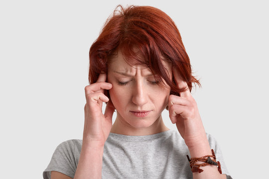 Headshot of stressful red haired young woman keeps both index fingers on temples, suffers from migraine, dressed casually, isolated over white background, tries to find solution. Headahe concept