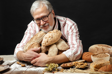 Open hearted old aged male Baker, loving his job so much, produces delicious organic bread and hugging it. Cooker is looking at camera and smiling while the bakery