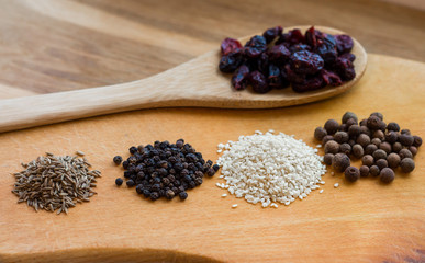 several handfuls of different spices (cumin, sesame, peppercorn) on a wooden table, wooden spoon is nearby