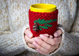 Girl in a light sweater holding a red cup of coffee with a knitted cover