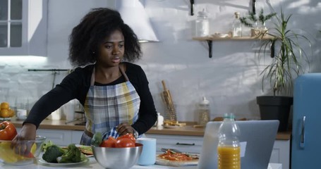 Medium shot of a young woman preparing food in the kitchen while watching a cooking tutorial on a laptop