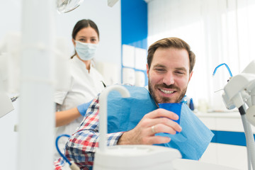 Obraz premium Attractive young man smiling at the dentist office, getting teeth repair, dental implants and teeth whitening by female dentist in a modern dental clinic. Male patient smiling in dentist chair.