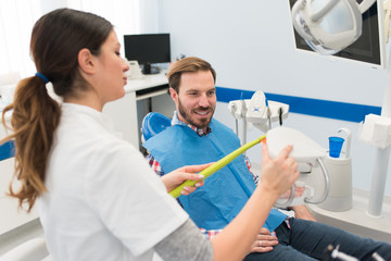 Obraz premium Young attractive happy smiling man at the dentist office, male patient. Female dentist holding toothbrush and teeth denture model. Stomatology Teeth whitening concept.