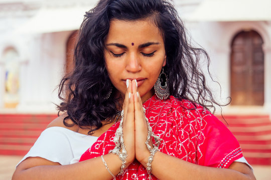 Young Indian woman in traditional sari red dress praying in a hindu temple goa india Hinduism.girl performing namaste gesture catholicism Delhi Street holi festival.om yoga meditation female model