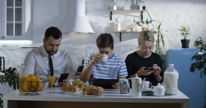 Medium shot of a family sitting side by side at the kitchen table and watching their smartphones and tablets