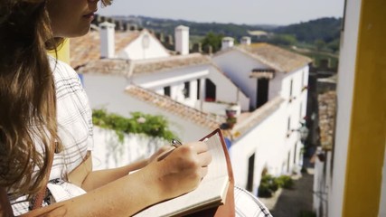 Unrecognizable female making notes in diary while sitting near building on sunny day in Obidos, Portugal