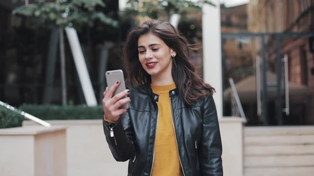 Portrait of happy young woman talking on video calling on the smartphone standing near building on the street