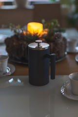 ein festlich gedeckter Esstisch mit Kerze und Gesteck zur Weihnachtszeit. Geschirr rahmt das Bild. Eine French Press steht im Fokus.