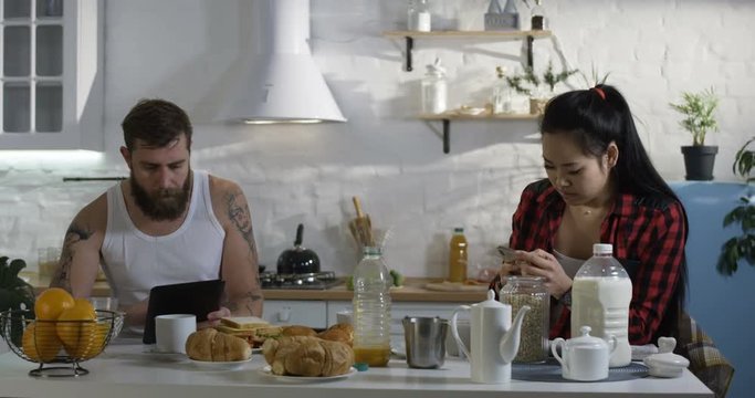 Medium shot of a couple busy using smartphone and tablet in the kitchen during breakfast