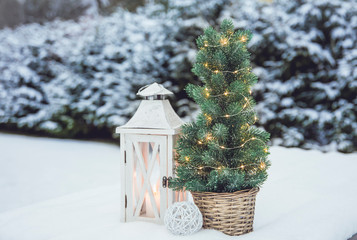 White wooden lantern with white wax candle lit and small Christmas fir tree with micro led lights in brown rattan flower pot in snow, snowy fir trees on the background, outdoors in winter.