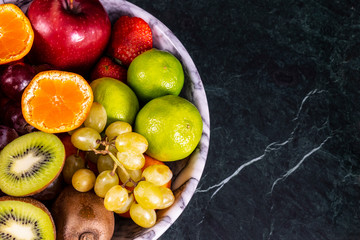 Fresh tropical fruitson in bowl on black marble background. Pineapple, papaya, mango, kiwi, grape, garnet, tangerines, bananas, apples, watermelon slices, pitaya or dragon fruit