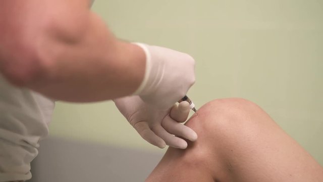 A close-up of the knee, hands in medicine gloves making an injection. Then they take the syringe out, put a sponge on the knee, bend, unbend it. Intra-articular injection of hyaluronic acid.