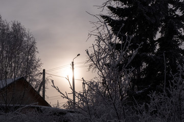 Winter evening in the snow-covered village. The roof of the house and a pillar with a lantern among the trees covered with hoarfrost. Theme of winter weather, lowering the temperature. Copy space.