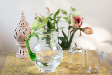 Glass pitcher with water and drinking glasses surrounded by flowers and peaceful elements.