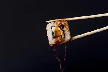Foto op Plexiglas sappig broodje tussen eetstokjes op zwarte achtergrond, druppels sojasaus druipend van sushi, voedselachtergrond, Japanse keuken © fantom_rd