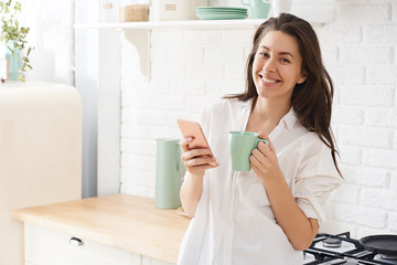Young woman using smartphone leaning at kitchen table with coffee mug and organizer in a modern home. Smiling woman reading phone message. Brunette happy girl typing a text message.
