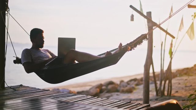 Young businessman working on his computer on vacation on a tropical island in the rays of the setting sun. A freelancer works on travel.