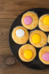 Muffins with pink and yellow cream, on rustic background. Black plate. Top view.