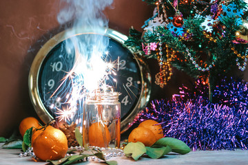 Fototapeta na wymiar Christmas composition consisting of hours, the arrow of which is at 12 o'clock, glasses of champagne with bubbles, sparklers in the bottlel, decorated Christmas tree, tangerines and other decorations
