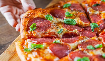 woman Hand takes a slice of Pepperoni Pizza with Mozzarella cheese, salami, Tomatoes, pepper, Spices and Fresh Basil. Italian pizza on wooden table background
