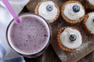 High glass of blueberry smoothie is next to tartlets filled with curd cream and decorated with blueberries on top are on a wooden board, top view