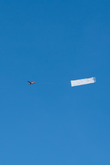 A small red airplane is pulling a white banner in the blue sky.